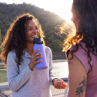 Zwei Frauen lächeln und unterhalten sich im Freien, eine davon hält eine Hydro Flask® 32 oz (946 ml) Weithalsflasche mit flexiblem Strohhalmverschluss in der Hand.