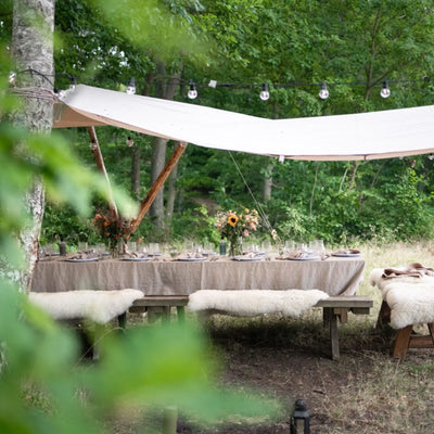 Essplatz im Freien in einem Wald mit einem Tisch unter einem weißen Nordisk® Kari 20 Tarp und Lichterketten.