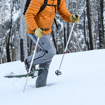 Eine Person in einer leuchtend orangefarbenen Jacke mit MSR® Lightning™ Trails - Schneeschuhen auf einem verschneiten Weg in einem Wald.