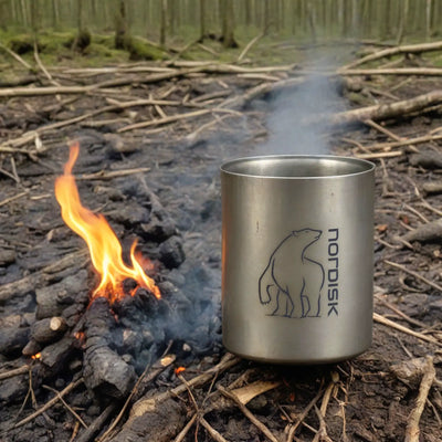 Ein Nordisk® Titanium Becher 450ml – Doppelwandiger Thermobecher mit Logo neben einem kleinen Lagerfeuer auf dem Boden platziert.