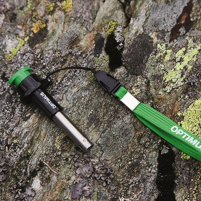 Ein Optimus Sparky Piezo Anzünder-Feueranzünder, Teil eines Optimus-Outdoor-Kits, mit einem grünen Griff, der an einem schwarzen Schlüsselband befestigt ist, der auf einem mit Flechten bedeckten Felsen ruht.