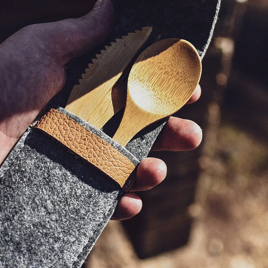 Eine Hand hält eine graue Filztasche mit einem ØYO Tourbesteck Bambus Besteck 3-teilig und einem Kamm mit natürlichem Licht, das Schatten wirft.