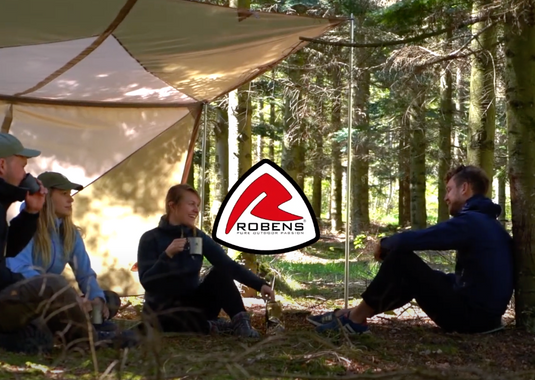 Eine Gruppe von Freunden unterhält sich unter einem Blätterdach im Wald, wobei das Robens-Logo sichtbar ist.