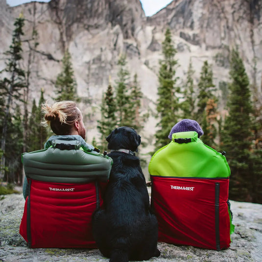 Zwei Personen und ein Hund sitzen in Therm-a-Rest® Trekker Chairs und genießen die Aussicht auf eine Berglandschaft.