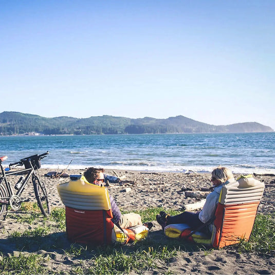 Zwei Personen entspannen sich in Therm-a-Rest® Trekker Chairs am Strand, während ein Fahrrad in der Nähe geparkt ist, und genießen den Blick auf das Meer und die Hügel in der Ferne.