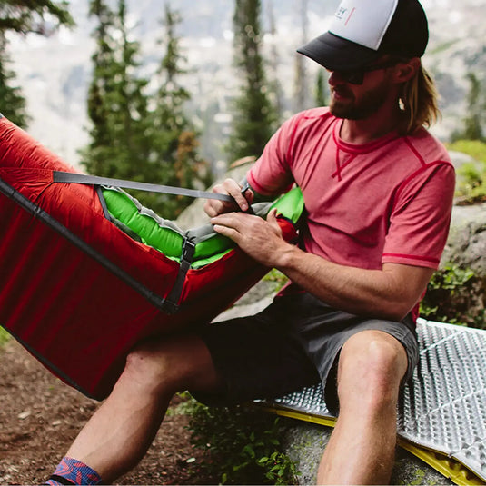 Mann passt Campingausrüstung an, darunter einen Therm-a-Rest® Trekker Chair, während er im Wald sitzt.