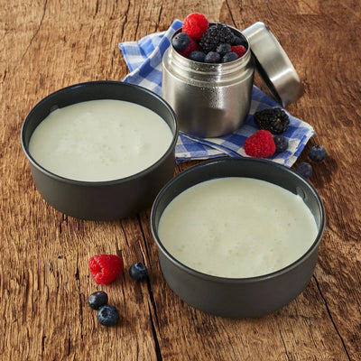 Zwei Schüsseln Joghurt mit frischen Beeren und Trek'n Eat Vollmilchpulver 2 Liter auf einer Holzoberfläche neben einem kleinen Metallbehälter.