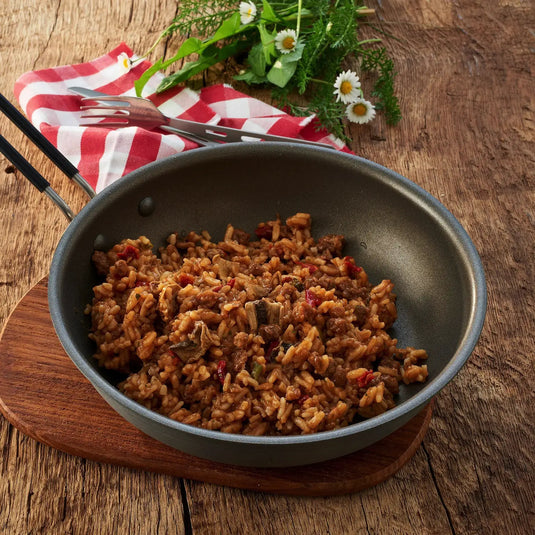 Eine Pfanne gekochtes Trek'n Eat Rindfleisch Stroganoff mit Reis auf einem Holztisch mit einer karierten Serviette und Wildblumen daneben.