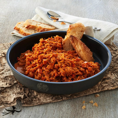 Eine Pfanne Trek'n Eat Chicken Tikka Massala mit exotischen Gewürzen und Kräutern, dazu ein Stück Brot, auf einer Holzunterlage.