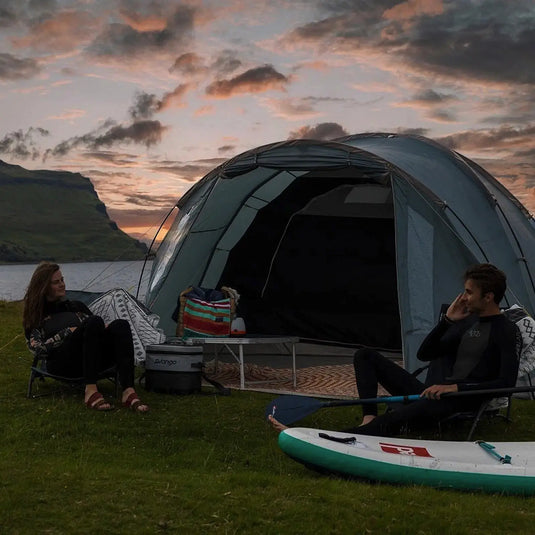 Zwei Personen entspannen sich neben einem Vango™ „Cragmor 400“ Kuppelzelt, 4-Personen mit einem Paddleboard in der Nähe, vor einer Hügelkulisse in der Abenddämmerung.