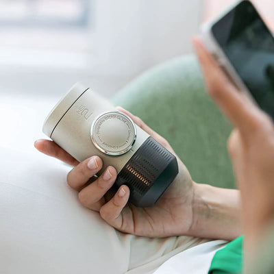 Eine Person hält eine WACACO® Minipresso NS2 mit einer für Espresso Kapseln konzipierten Wählschnittstelle, möglicherweise ein Smart-Home-Gadget, während sie ein Smartphone benutzt.