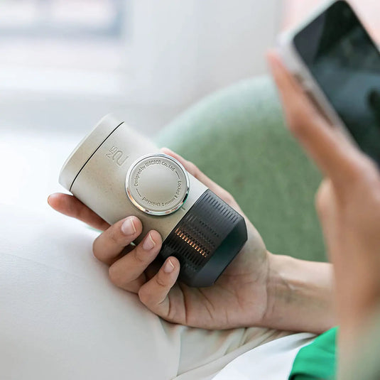 Eine Person hält eine WACACO® Minipresso NS2 mit einer für Espresso Kapseln konzipierten Wählschnittstelle, möglicherweise ein Smart-Home-Gadget, während sie ein Smartphone benutzt.