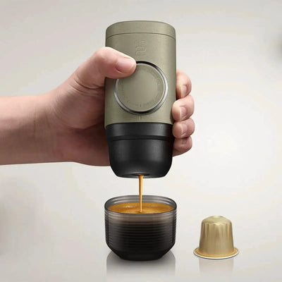 Eine Hand, die eine WACACO® Minipresso NS2 hält, während sie Espresso in eine kleine Tasse ausgibt, mit einer leeren Kapsel daneben.