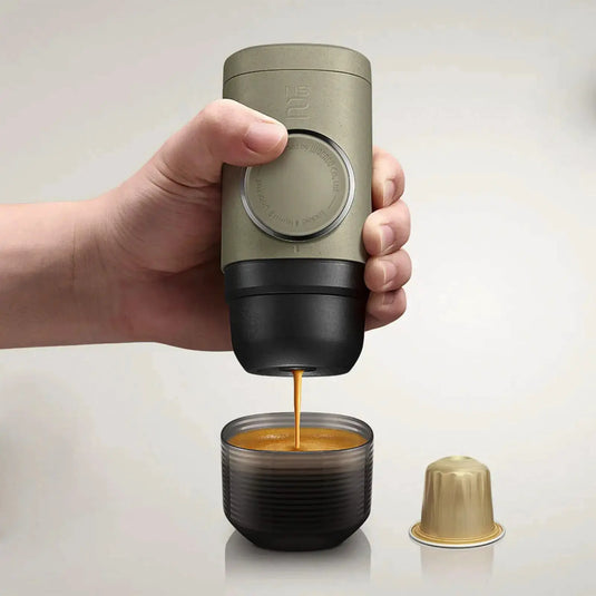 Eine Hand, die eine WACACO® Minipresso NS2 hält, während sie Espresso in eine kleine Tasse ausgibt, mit einer leeren Kapsel daneben.